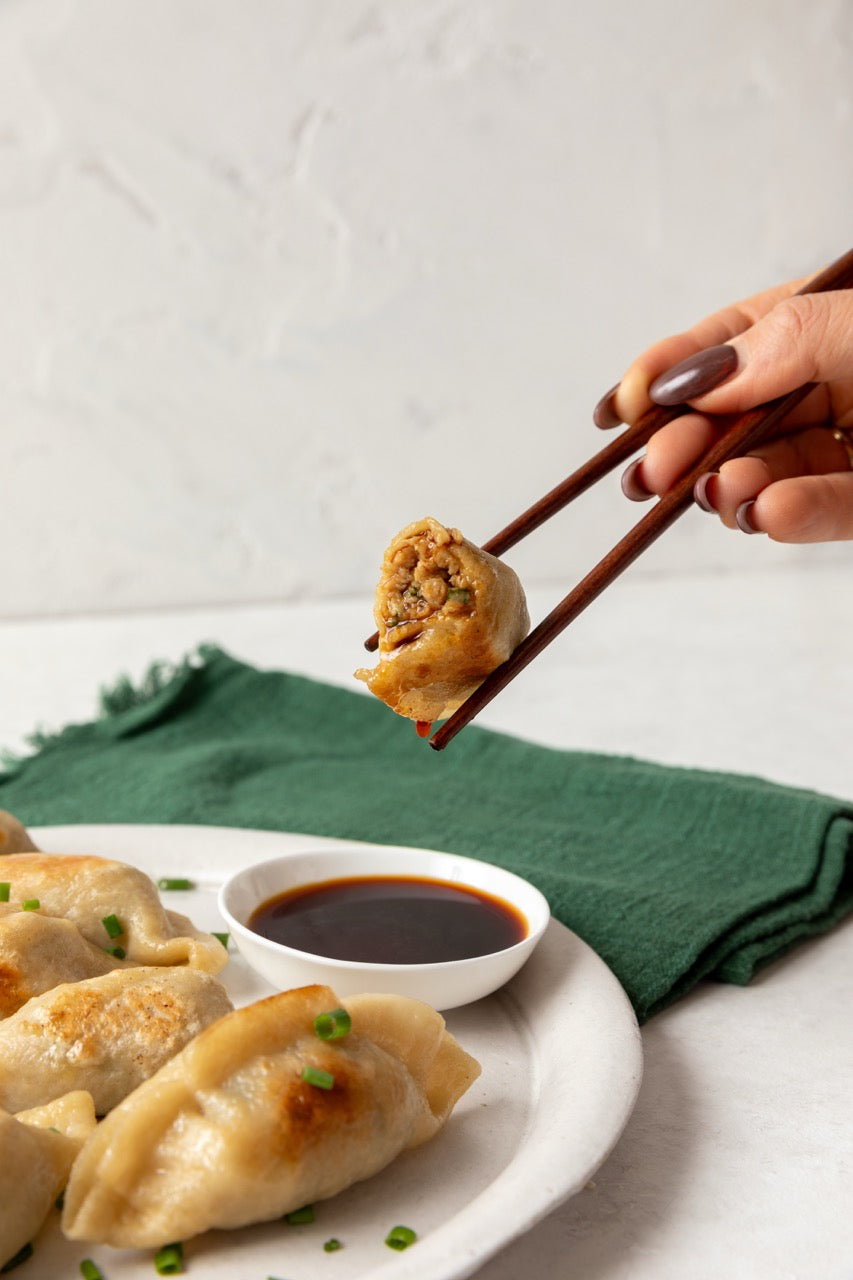 Person holding a FroPro High Protein dumpling with chopsticks over a plate of FroPro high protein dumplings and a bowl of sauce.