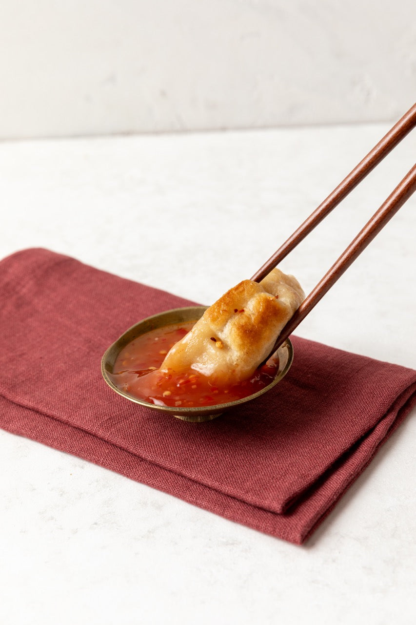 FroPro high protein Dumpling being held with chopsticks over a small bowl of red sauce on a red cloth.
